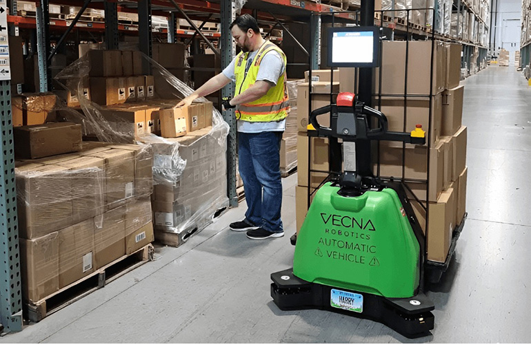A man standing next to a green Vecna Robotics pallet handling robot, part of the company's new CaseFlow system, and scanning boxes.