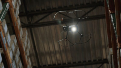 A Gather AI drone flying above head in a warehouse, with some shelving seen in the background on the sides of the image. Bosma uses Gather AI's drones, AI, and accessible interface.