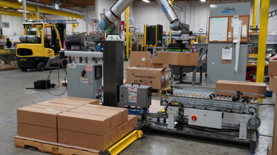 A Columbia/Okura miniPAL cobot system stacking boxes from a conveyor belt onto a pallet.
