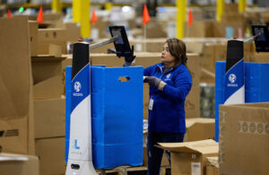 A female GEODIS employee wearing blue performing a pick with the help of a LocusBot in a warehouse setting.