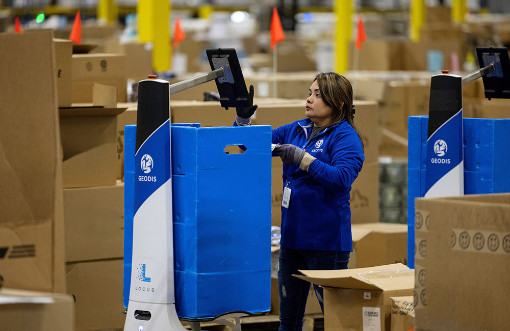 A female GEODIS employee wearing blue performing a pick with the help of a LocusBot in a warehouse setting.