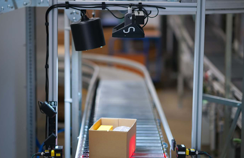 A box with two objects in it moving down a conveyor belt  under a station with Sereact vision technology. 